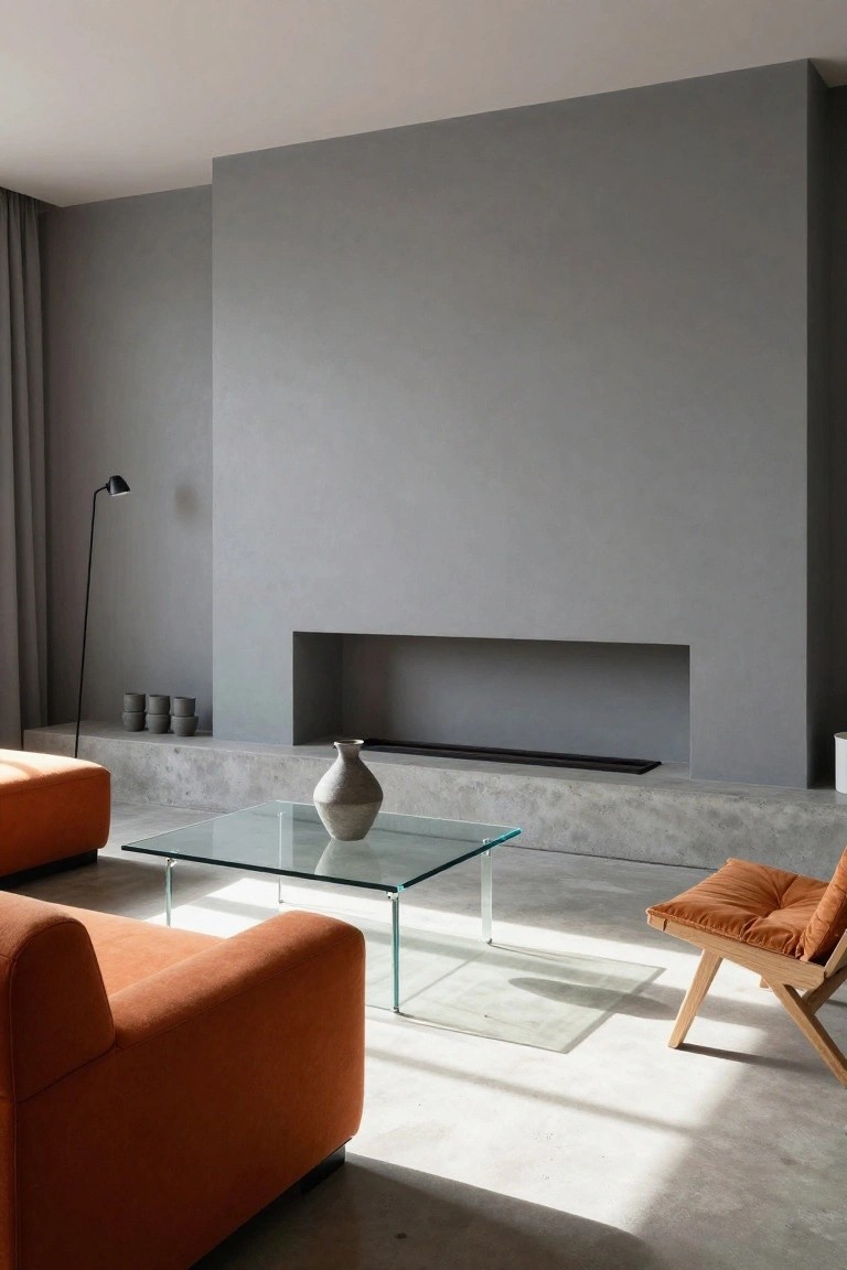 Modern living room with two burnt orange fabric sofas flanking a clear glass coffee table on polished concrete floor, large gray wall with recessed fireplace, tall floor lamp, wooden chair, and ceramic vases.