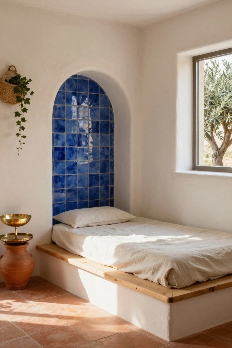 Bedroom with low wooden platform bed on elevated white base and white linens, backed by blue tiled arched wall, hanging ivy basket, gold bowl on terracotta pot stand, window overlooking olive tree, terracotta tile floor.
