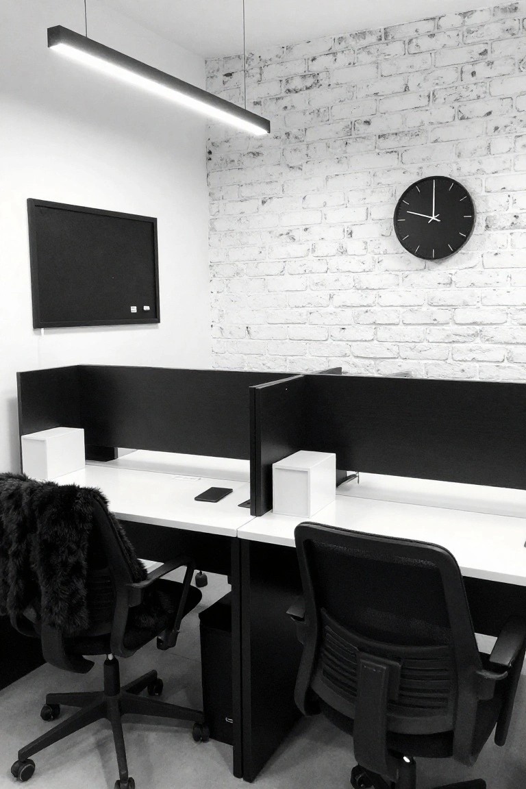 Black and white photo of two white desks separated by tall black partitions in a small office with black chairs, white brick wall, wall clock, and bulletin board.