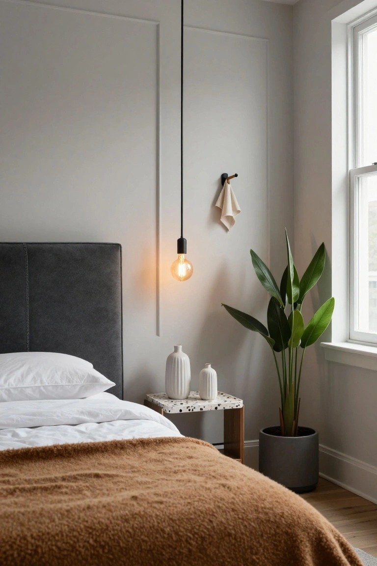 Corner of a bedroom with a gray upholstered headboard bed topped with white pillows and sheets under a brown throw blanket, next to a speckled side table holding two white vases, a tall potted plant in a gray pot, and a black pendant light with exposed bulb hanging from the ceiling.