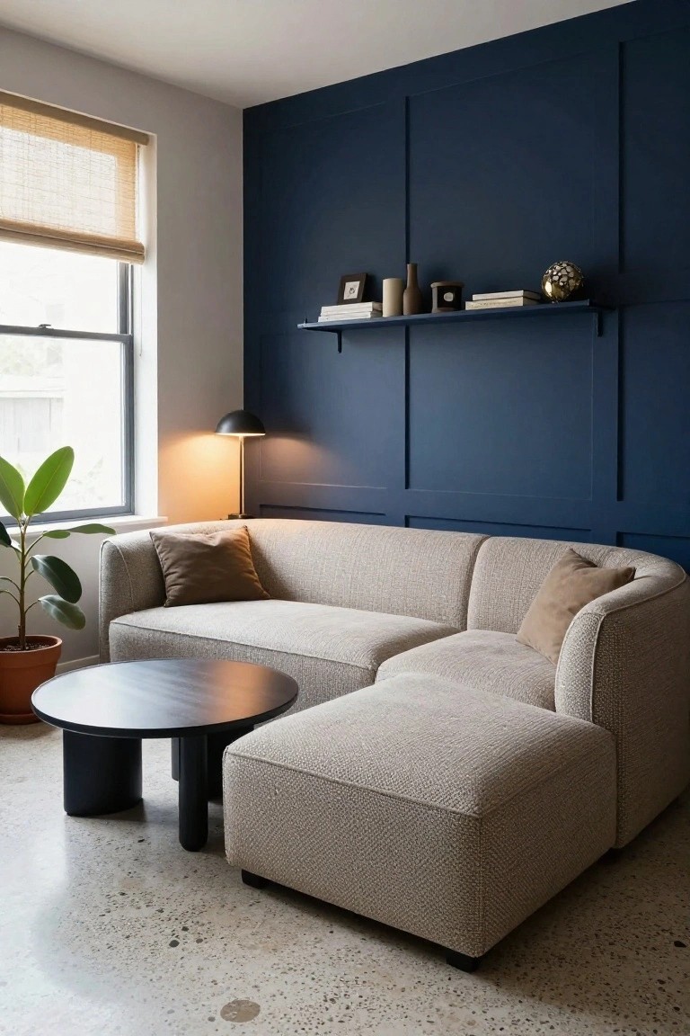 Modern living room corner with light beige curved sectional sofa, matching ottoman, black round coffee table, potted plant, desk lamp, and floating shelves on navy paneled wall.
