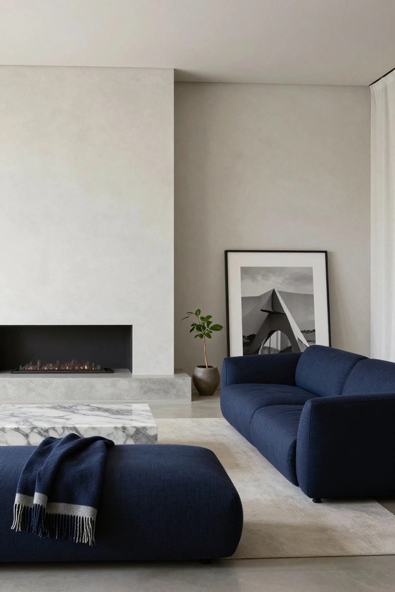 Modern living room with L-shaped navy blue sofa, matching blue ottoman with navy throw blanket, white marble coffee table, linear gas fireplace in textured white wall, potted plant in ceramic pot, large black-and-white framed photo of architecture, and light beige rug on concrete floor.