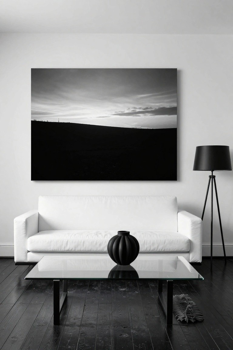 Modern living room featuring a white sofa against a white wall, large framed black-and-white landscape photograph above it, glass coffee table with black vase, black tripod floor lamp, and dark wood floors.