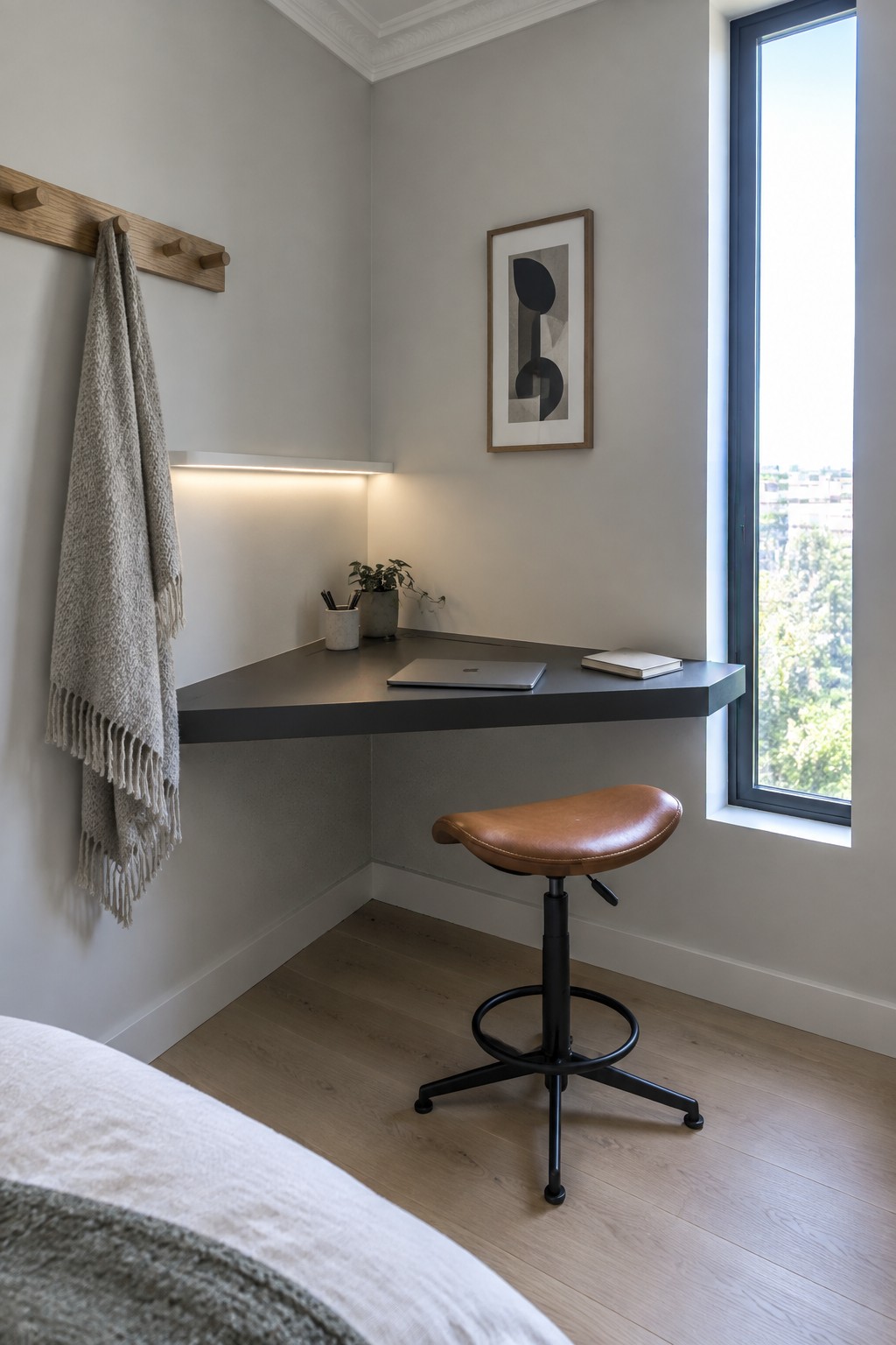 A bedroom corner with a floating dark desk, brown leather stool, laptop, and framed artwork on a light gray wall next to a window.