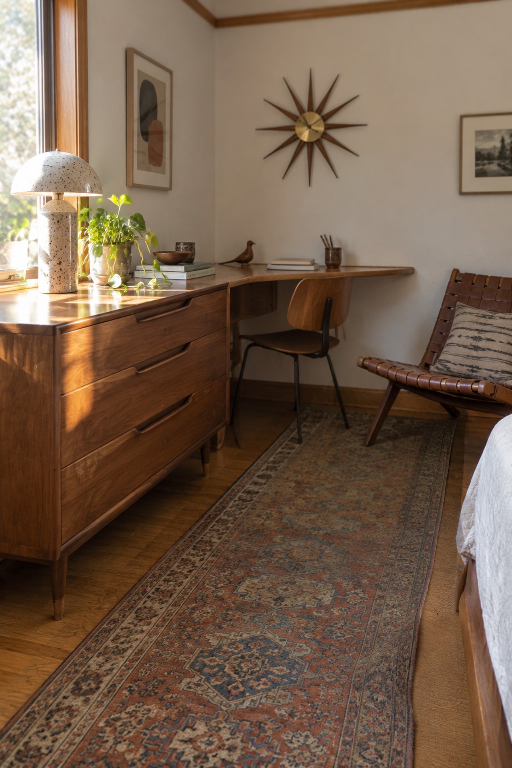 A bedroom corner featuring a wooden L-shaped desk with a matching chair, a wooden dresser with a lamp and plant, a starburst clock on the wall, and a long patterned rug on the hardwood floor.