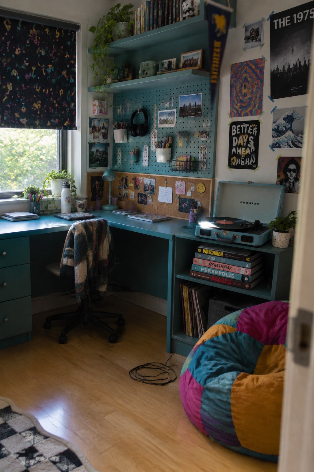 A teal corner desk with pegboard and shelves above it sits in a bedroom beside a window, with a record player on the right and a colorful beanbag chair in the foreground.