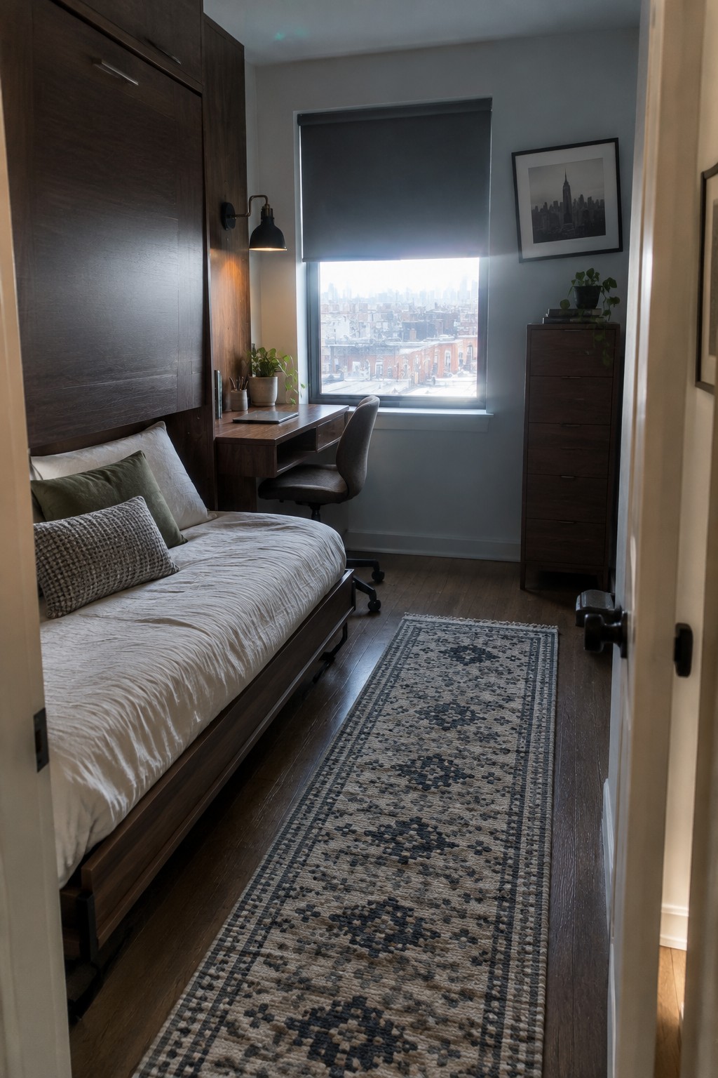Compact bedroom with bed, desk, office chair, city view window, wooden dresser, and patterned rug.