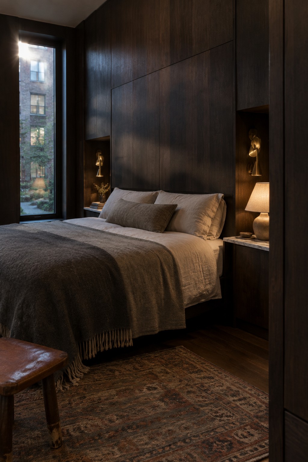 A bedroom interior with full-height dark wood paneling behind the bed, neutral bedding, wall sconces, and a large window on the left.