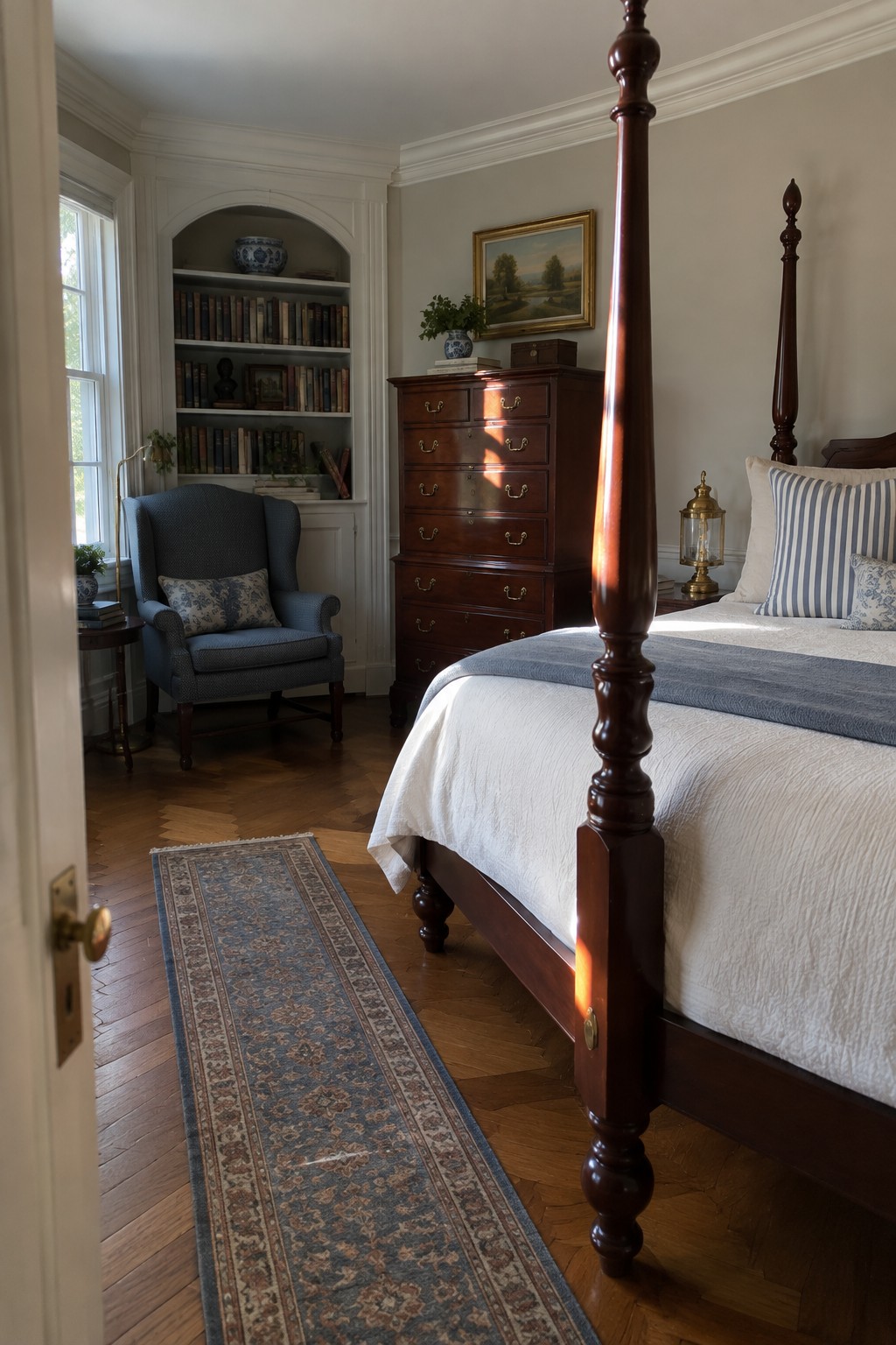 A bedroom featuring a dark wood four-poster bed with white and blue bedding, a matching tall dresser, built-in bookshelves, and a blue upholstered armchair on a wood floor with a patterned runner.