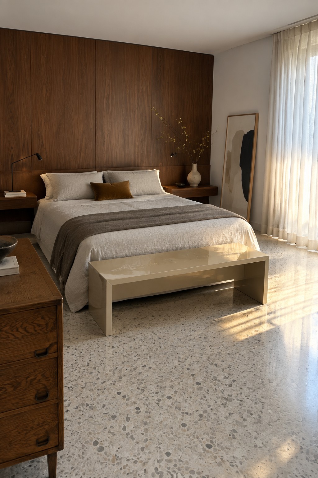 A modern bedroom featuring a floor-to-ceiling wood-paneled wall behind the bed, white and gray bedding, a glossy beige bench at the foot of the bed, and a wooden dresser in the foreground on a terrazzo floor.