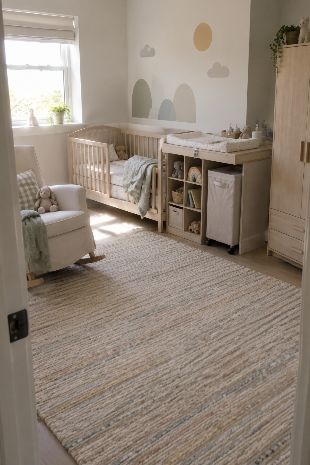 A bright nursery with a wooden crib, a freestanding changing table with storage bins and bins on wheels, a white rocking chair, and a woven striped rug.
