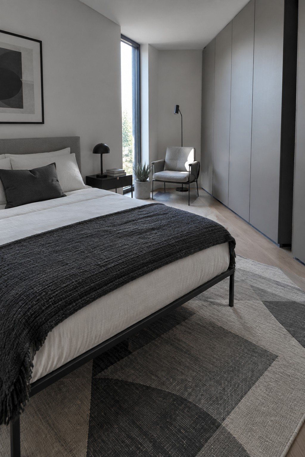 A modern bedroom with a bed that has a thin black metal frame, white bedding, and a dark textured throw, positioned on a patterned rug near a small black nightstand and a gray upholstered chair.