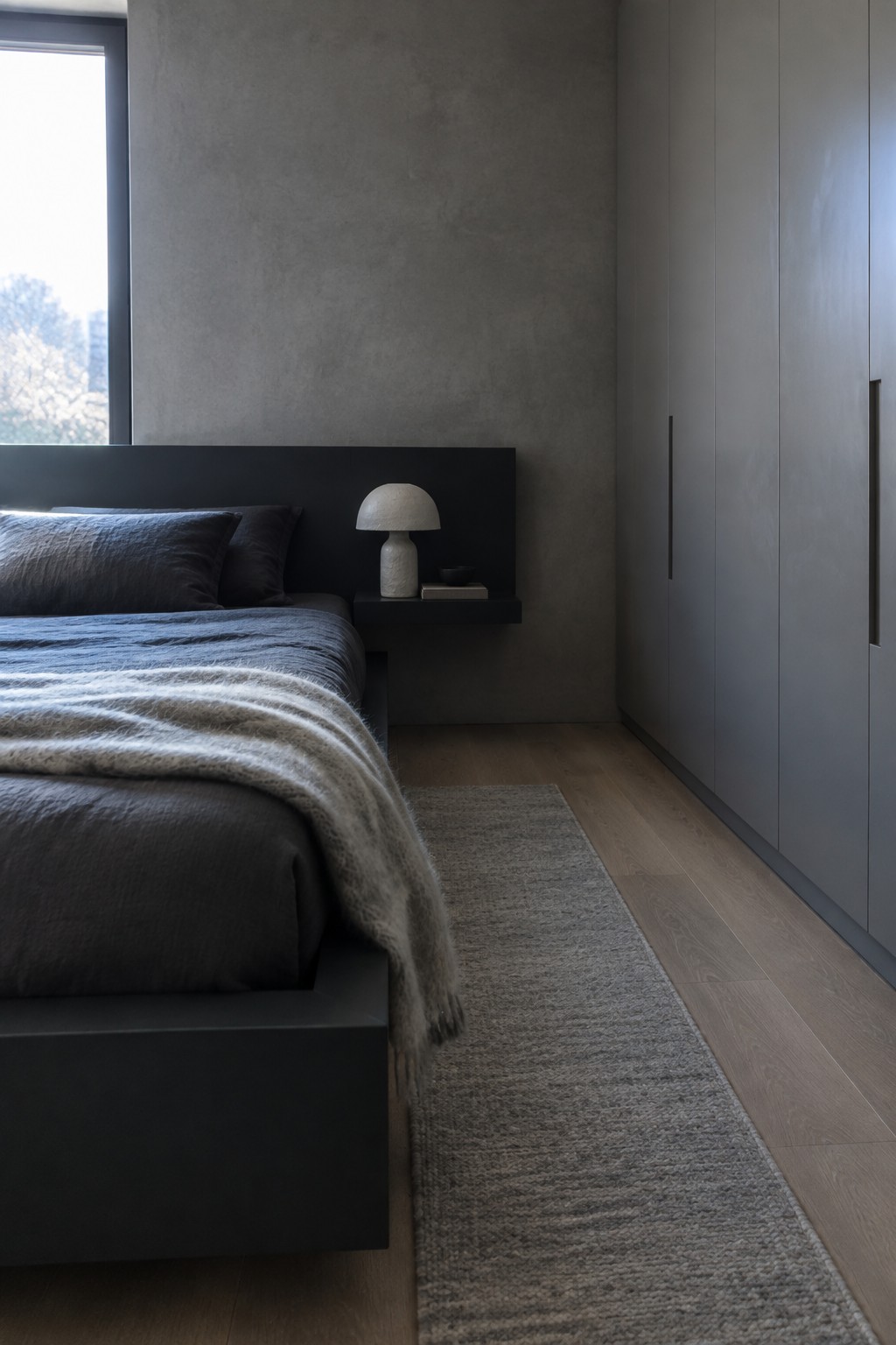 A modern bedroom featuring a black painted bed frame with dark linens, a floating black nightstand holding a small lamp, and tall gray cabinets along the right wall.