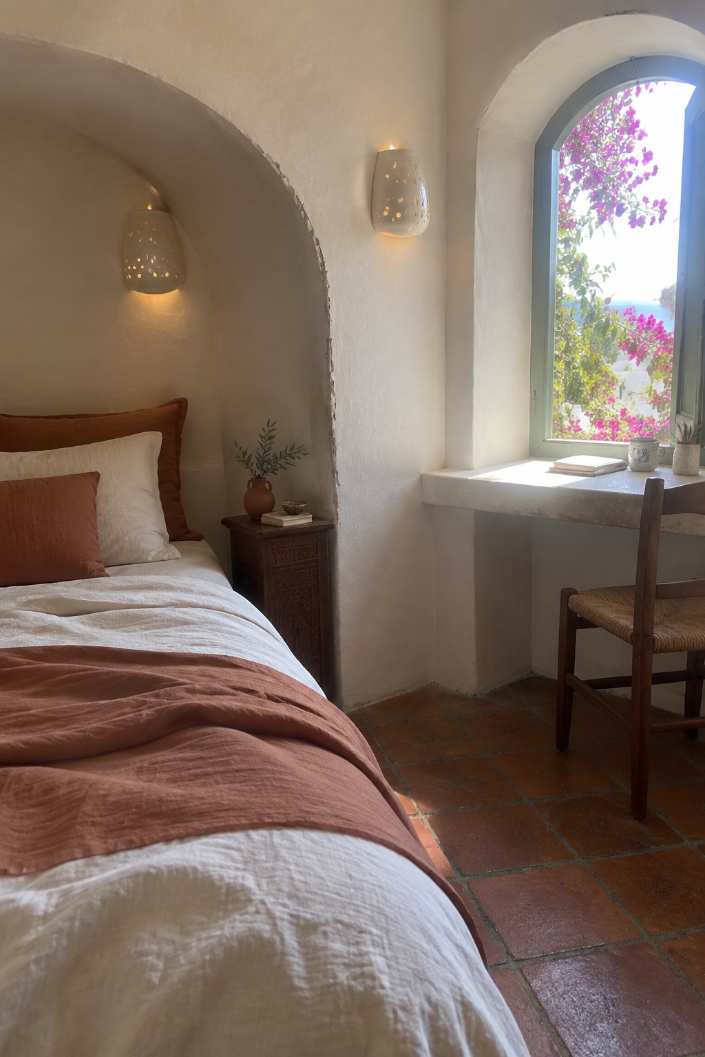 Cozy bedroom with terracotta linens, arched window overlooking sea, and wooden nightstand.