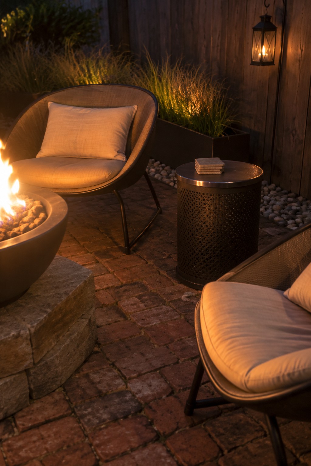 Two cushioned outdoor armchairs sit on a brick patio beside a round fire pit, with a metal side table and hanging lantern nearby.