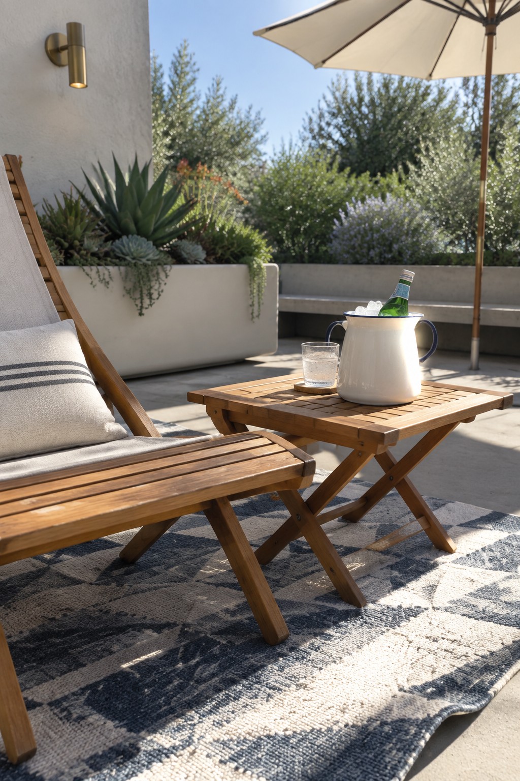 A wooden lounge chair and small folding table rest on a blue and white rug on a concrete patio, with an umbrella, ice bucket, and potted plants visible nearby.