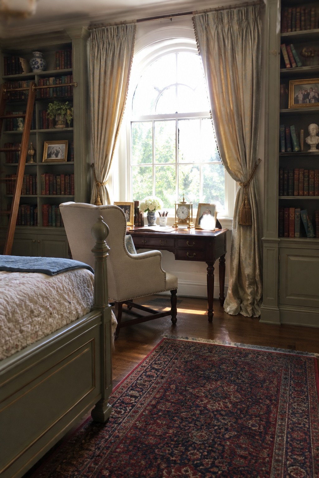 A bedroom interior showing a dark wood desk placed in front of an arched window, flanked by built-in sage green bookshelves filled with books, and a light upholstered armchair pulled up to the desk.