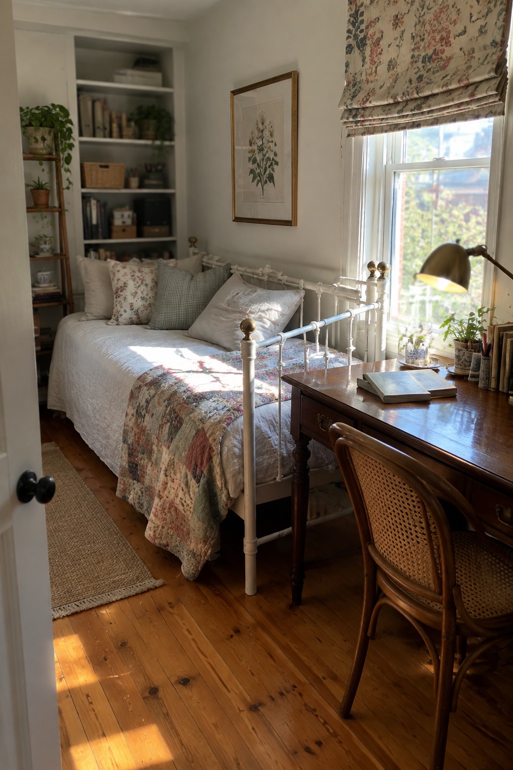 A bedroom interior showing a white metal bed with a patchwork quilt and a wooden desk with a cane chair placed beside a sunlit window.