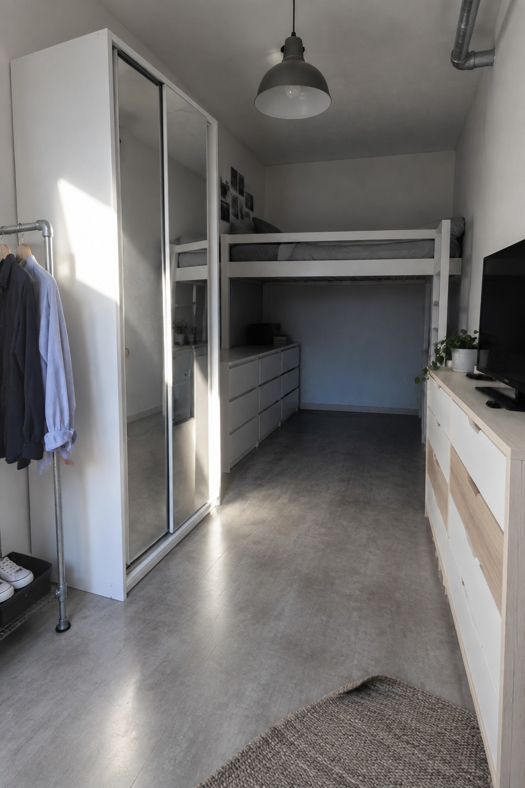 A compact bedroom with a white loft bed over built-in white drawers, a mirrored sliding-door wardrobe on the left wall, and a low dresser with a television on the right.