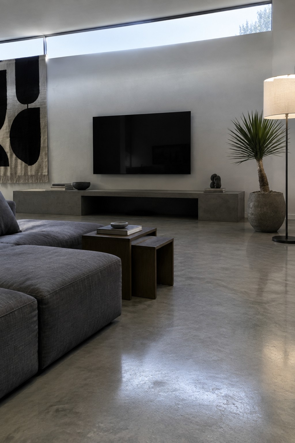 A modern living room featuring a wall-mounted flat screen TV above a long concrete media console, with a gray sectional sofa and wooden nesting tables in the foreground.