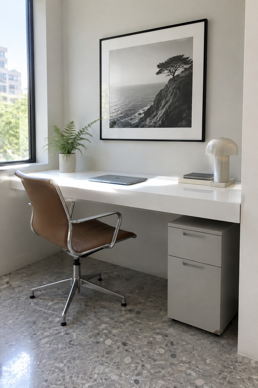 White minimalist desk with brown leather chair, laptop, mushroom lamp, plant, and framed coastal photo