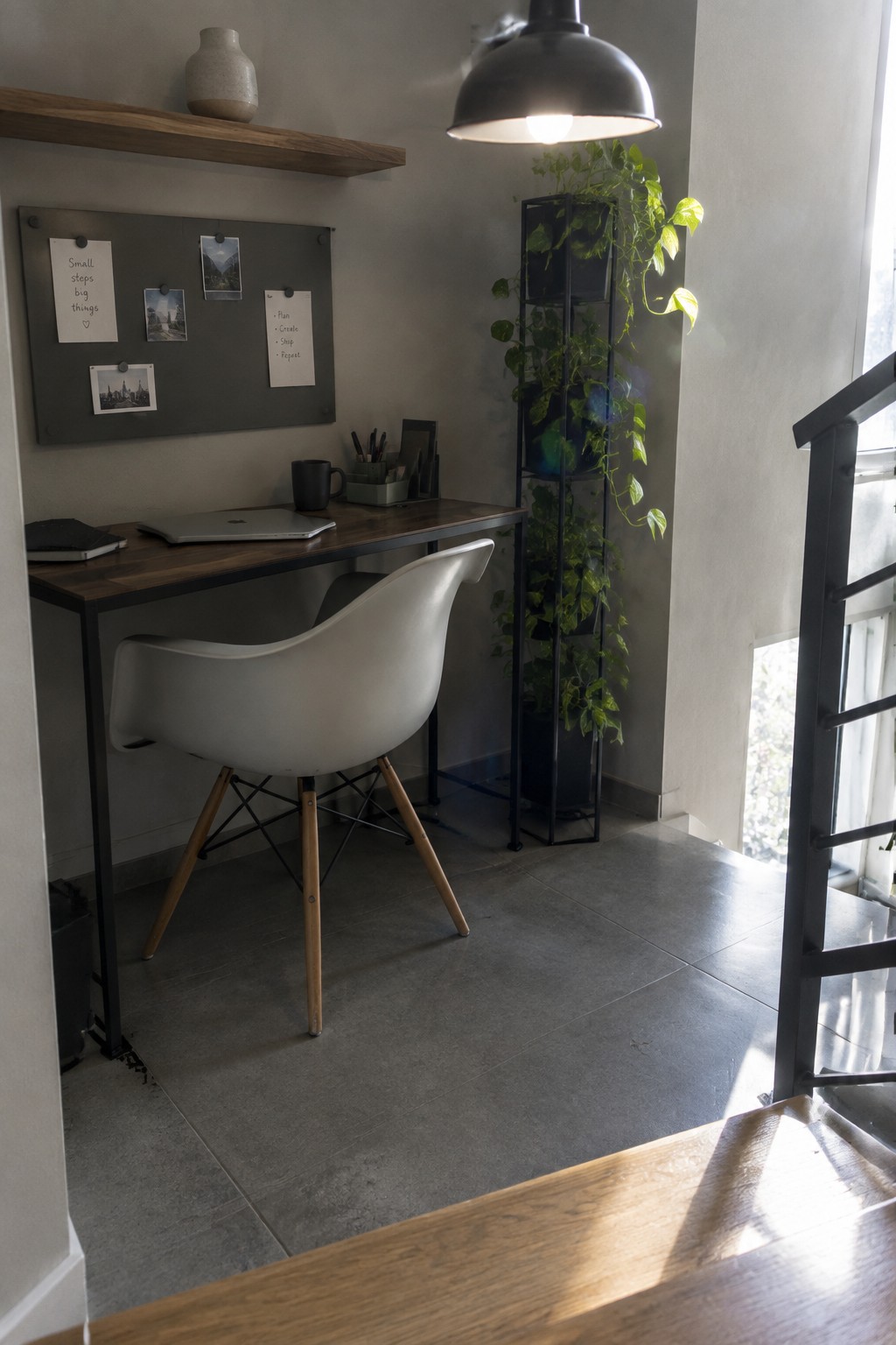 A compact wooden desk with a white molded chair sits under a gray wall-mounted notice board displaying notes and photos, with a tall black plant stand holding greenery nearby.