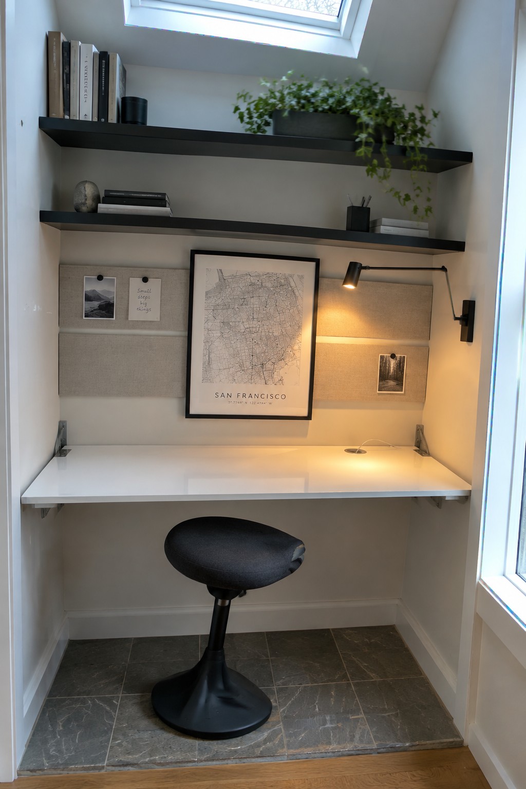 A compact home office nook featuring a white wall-mounted desk with black floating shelves above, fabric wall panels, a framed map print, a black adjustable wall lamp, and a black stool on gray tile flooring.