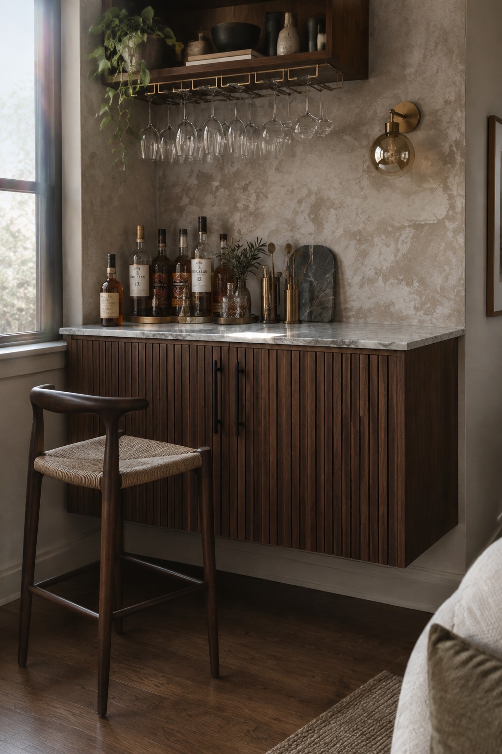 A dark wood bar cabinet with vertical slats and marble top sits below floating shelves holding wine glasses, with a wooden bar stool in front.