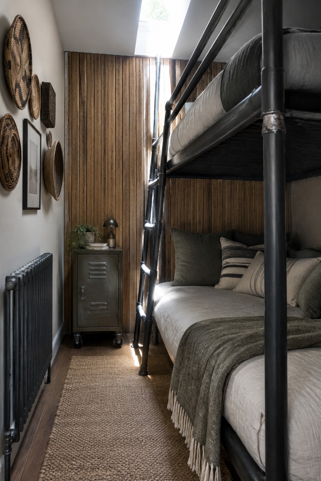 A narrow bedroom with a black metal bunk bed, wood slat walls, a metal rolling cabinet, woven wall baskets, and a jute rug on wood flooring.