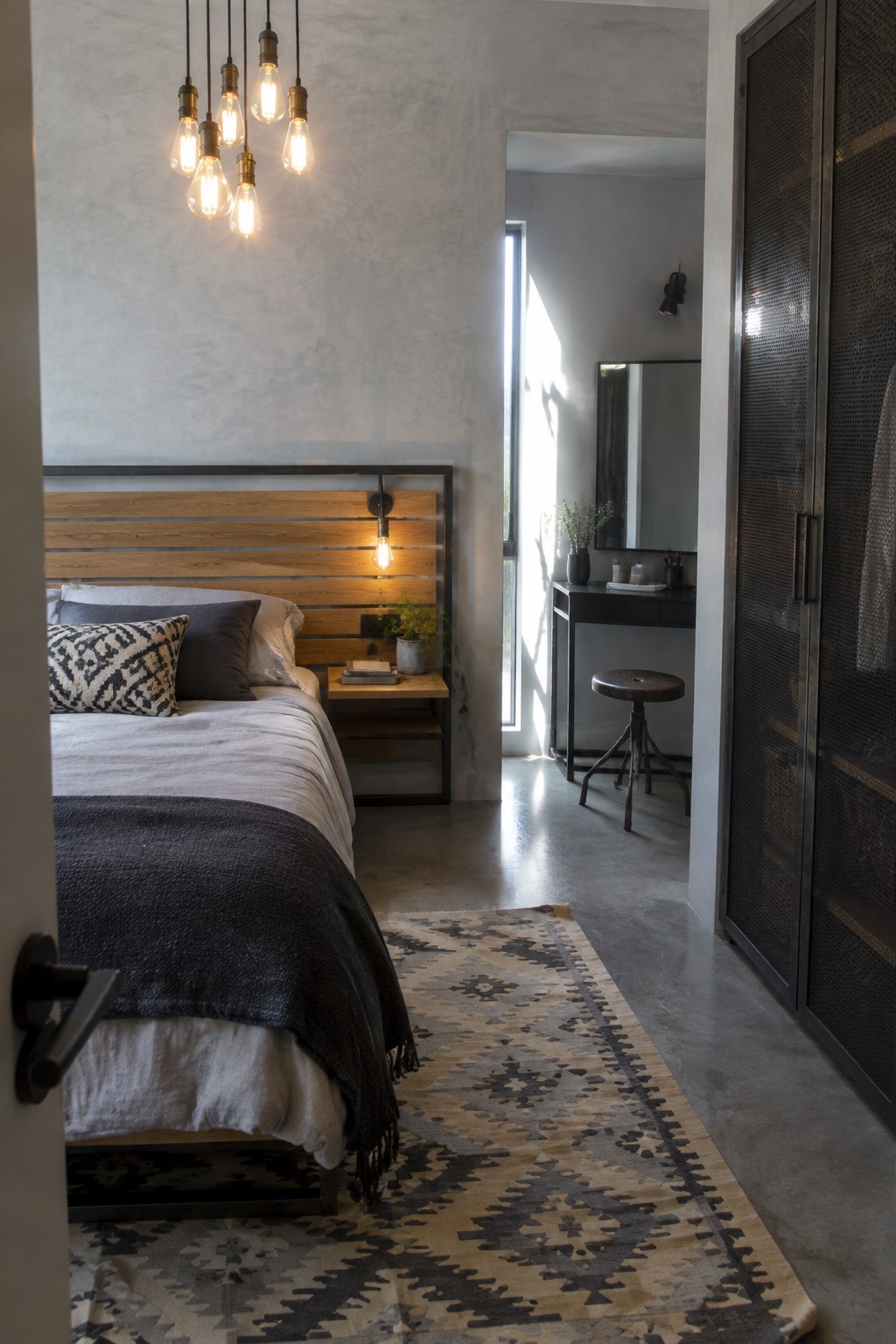 A bedroom with a bed featuring a horizontal wood slat headboard in a dark metal frame, several lit industrial pendant lights above, and a patterned rug on the concrete floor.