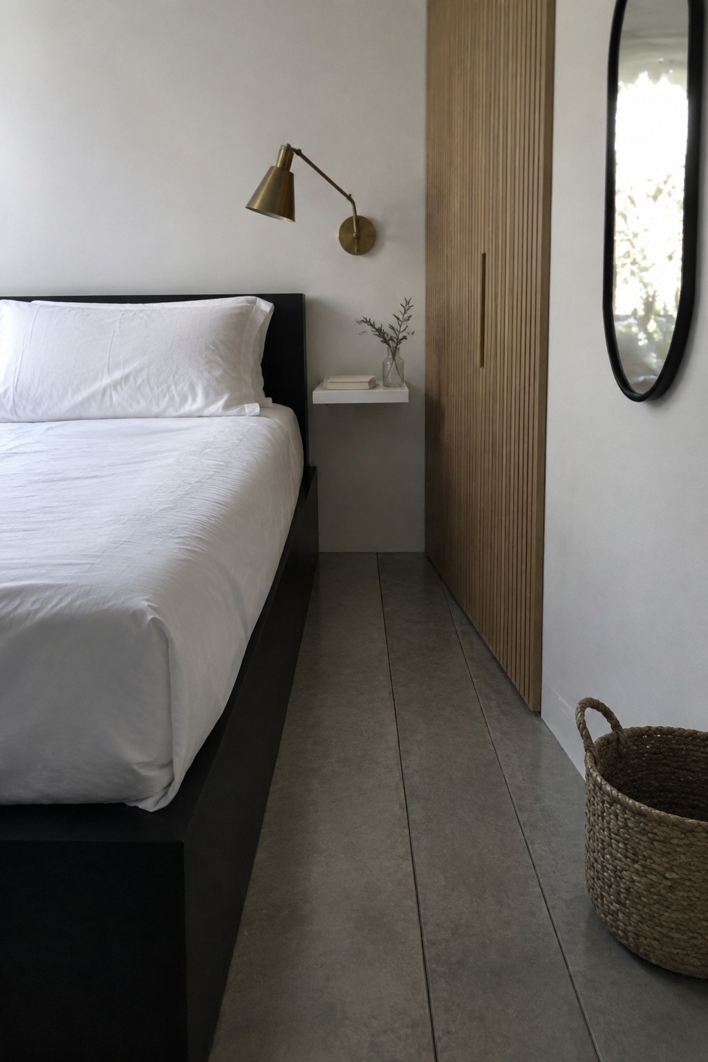 A minimalist bedroom featuring a bed with white bedding, a black platform frame, a floating white shelf with a small plant and book, a brass wall sconce, a vertical wood slat closet, and a woven basket on a concrete floor.