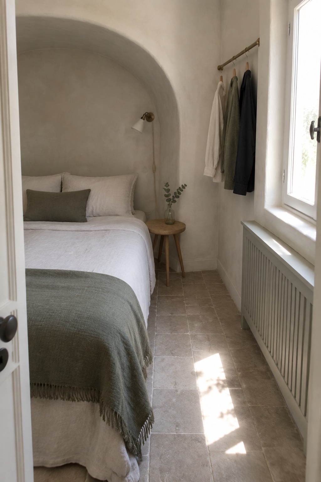 A small bedroom showing a bed with white and green linens placed inside a large arched alcove, with a narrow side area holding hanging clothes and a window seat.