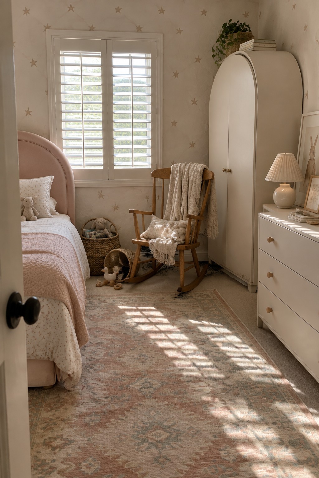 Cozy pastel nursery with pink bed, rocking chair, white wardrobe, and patterned rug.