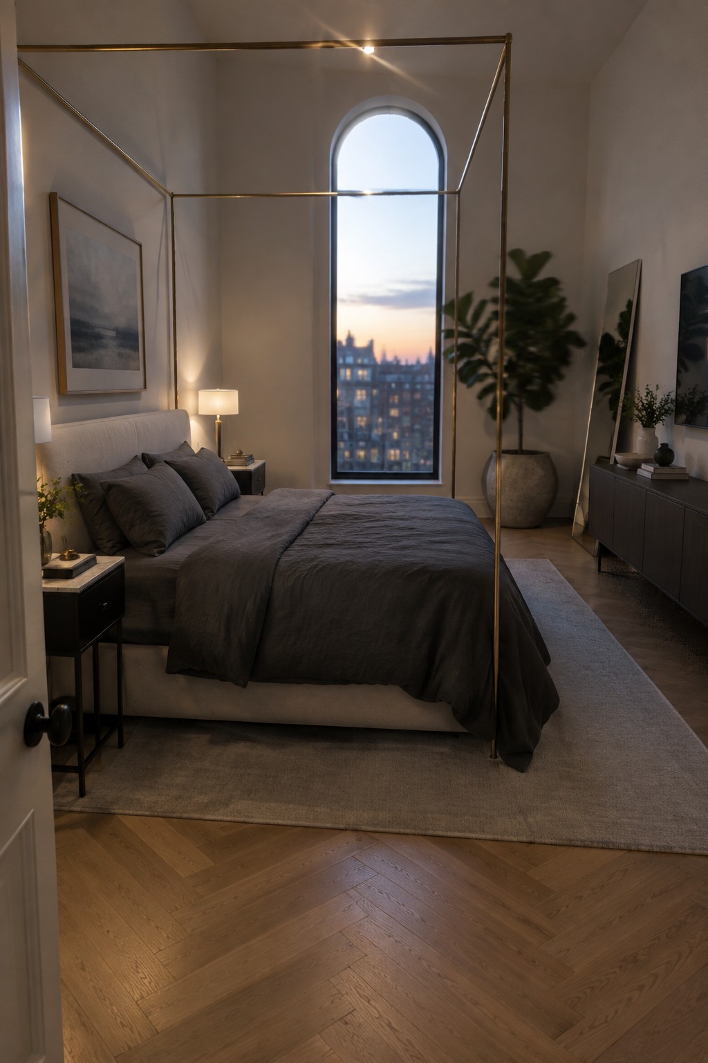 A modern bedroom featuring a thin brass canopy bed frame with dark gray bedding, placed in front of a large arched window overlooking city buildings at dusk.