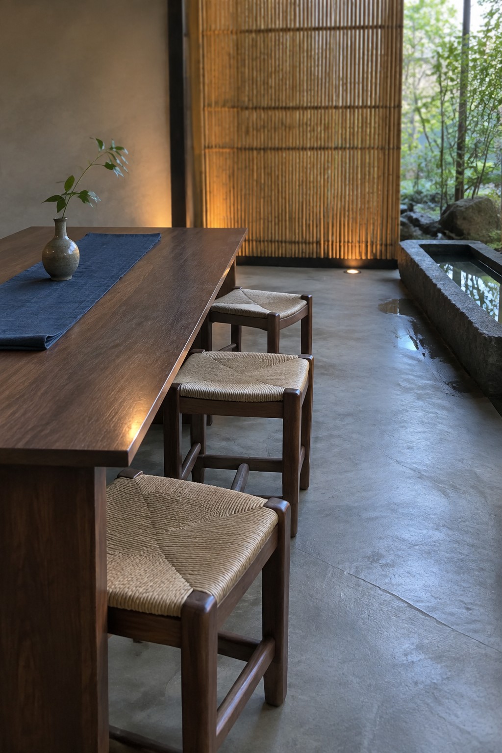A long wooden dining table lined with woven-seat wooden stools in a covered space next to a stone water feature.