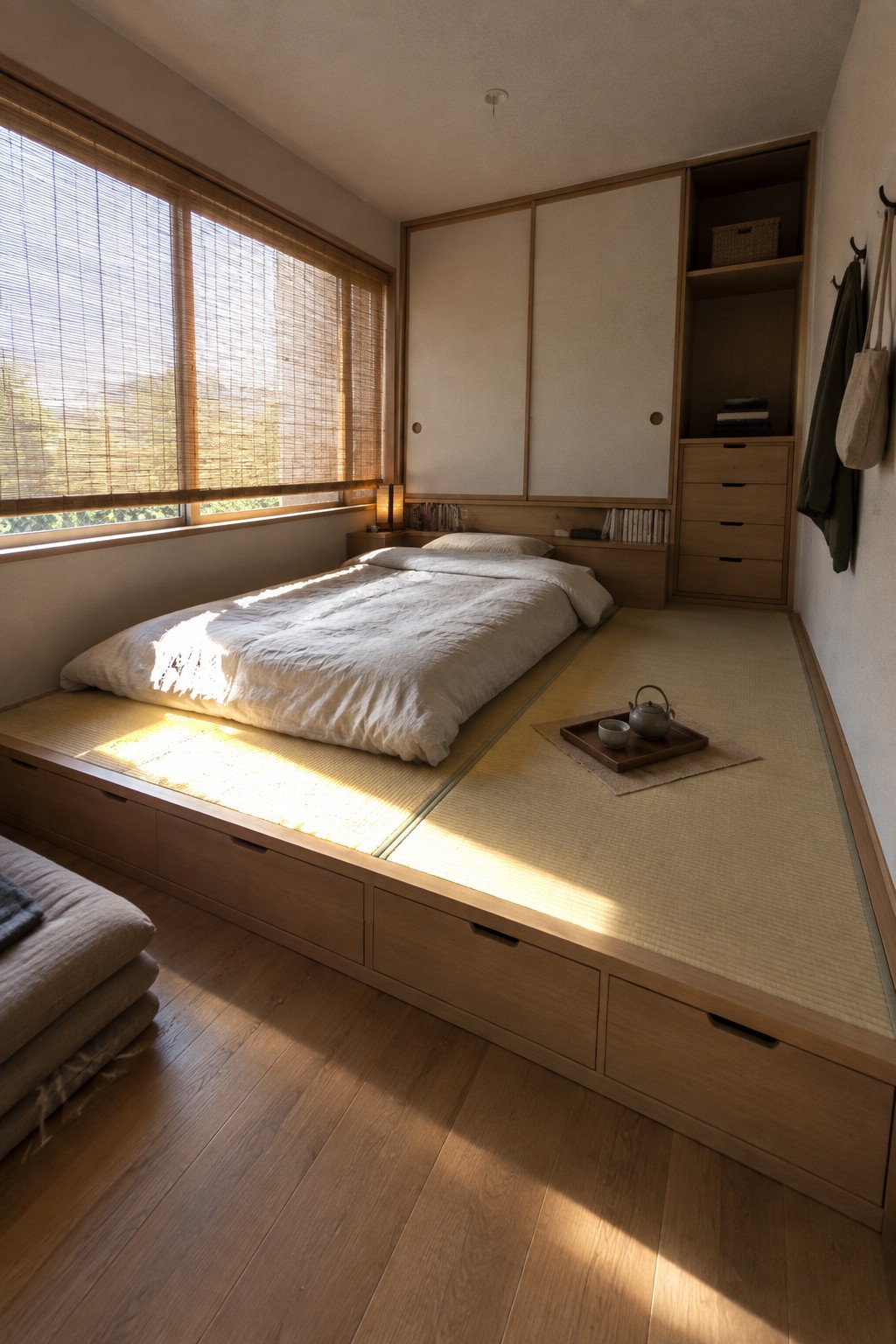 A minimalist bedroom featuring a raised wooden platform bed with tatami mats, white bedding, and multiple built-in storage drawers along the base, positioned beside a window with wooden blinds and adjacent wall cabinets.