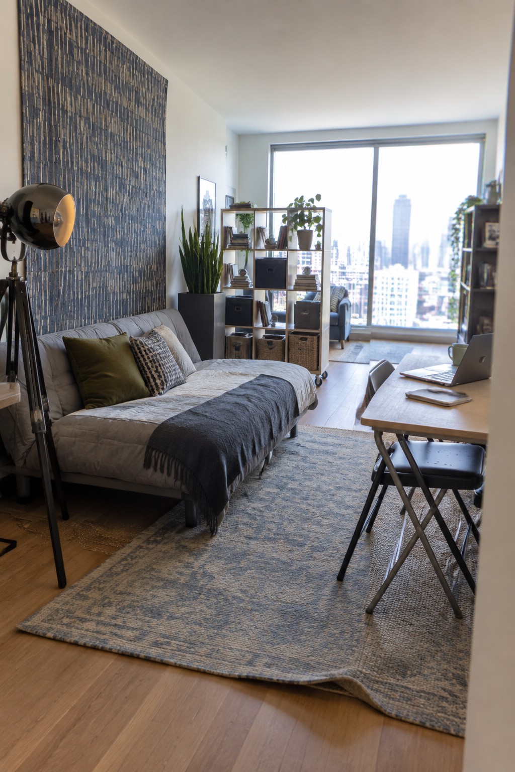 A living area with a gray upholstered daybed, patterned rug on wood floors, rolling white shelving unit, and large windows in the background.
