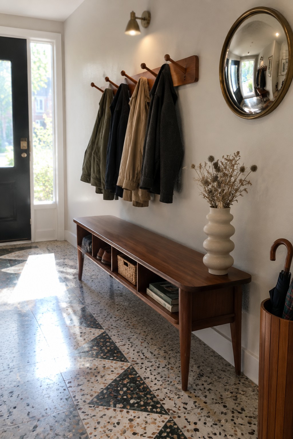 A narrow interior entryway featuring a long wooden bench with open cubby storage, a wall-mounted wooden coat rack holding several jackets, a white ceramic vase with dried flowers, and a round mirror above a terrazzo tile floor.