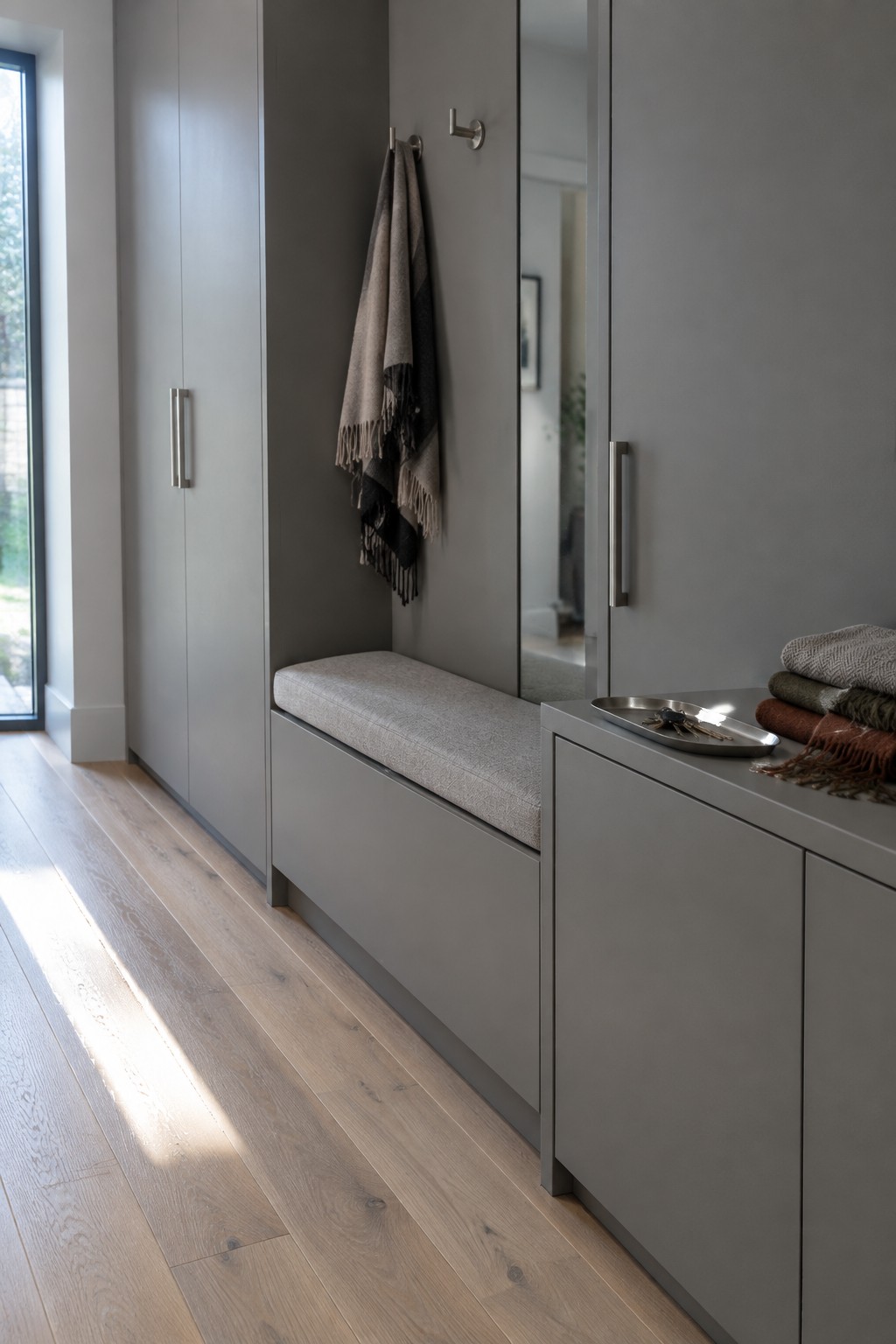A narrow interior entryway featuring gray built-in cabinets, a long cushioned bench, wall hooks with scarves, and a small countertop tray.