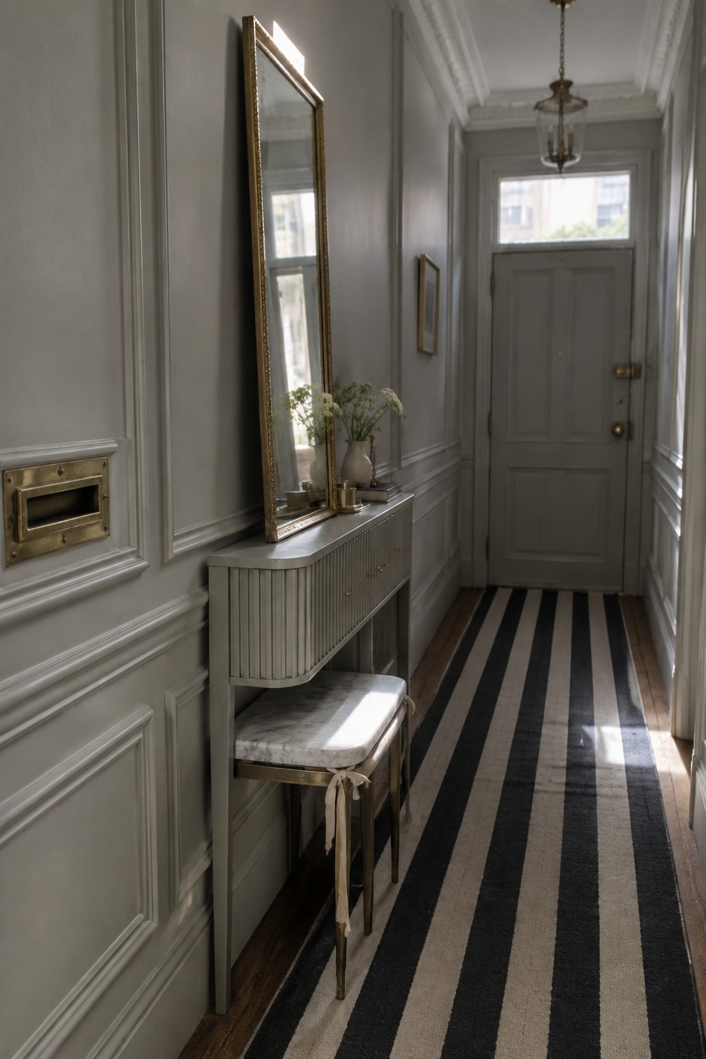 A narrow gray hallway features a slim console table with a marble-topped stool beneath a tall mirror, a black and white striped runner rug, and a gray front door at the end.