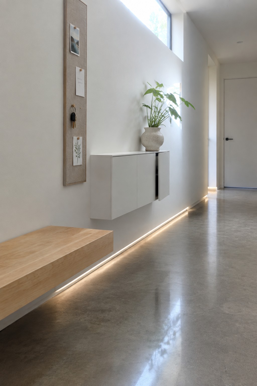 A narrow interior hallway with a floating light wood bench, white wall-mounted cabinet, and tall fabric pinboard on a white wall, with LED strip lighting along the base.