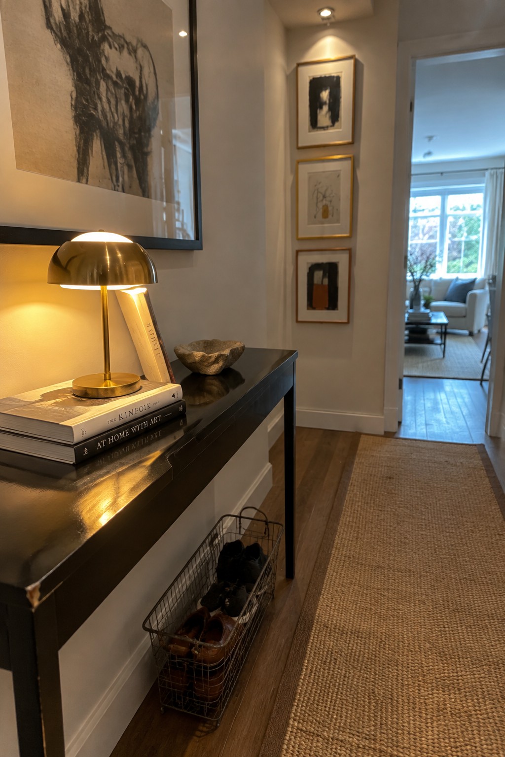 A narrow black console table in a hallway holds a lit brass lamp and stacked books, with a wire basket of shoes stored underneath on a wooden floor.