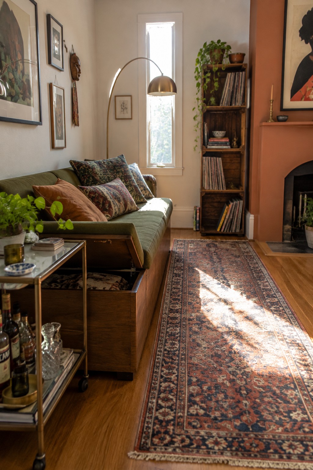 A living room with a green sofa that has an open lift-up storage compartment, a rolling metal cart with bottles and books, a long patterned runner rug, and a tall wooden bookshelf beside a fireplace.