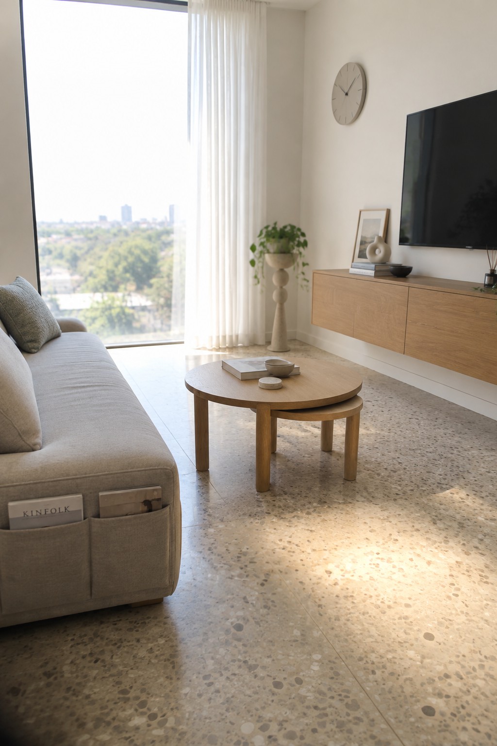 A bright living room featuring a light gray sofa with magazine pockets, round wooden nesting coffee tables, a wall-mounted television on a light wood console, and a large window with white curtains.